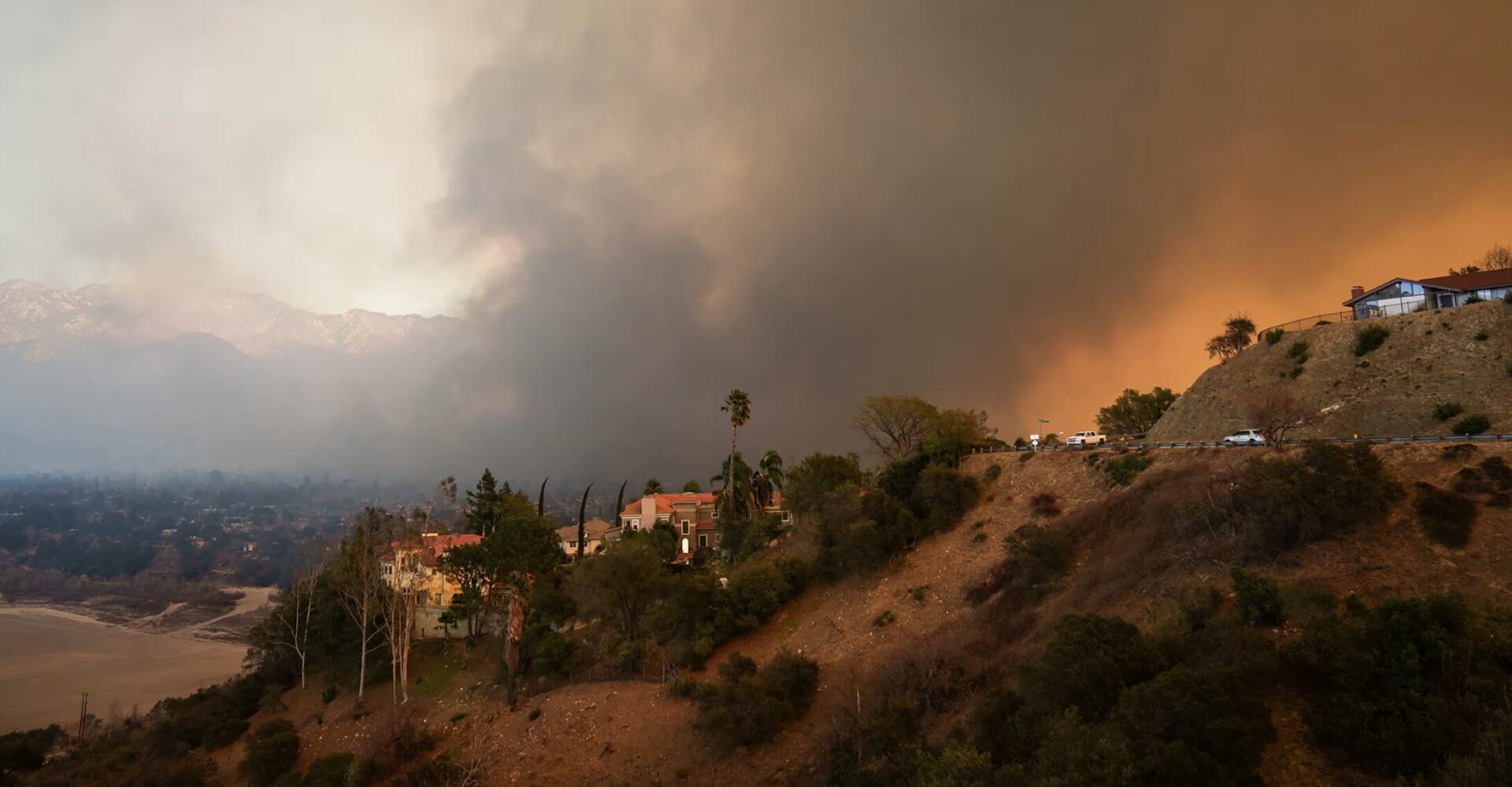 A hillside with houses and palm trees under a thick, smoky sky. Smoke from a fire dominates the right side, blending with the orange glow of flames. The left side shows a hazy, distant mountain range under a lighter sky.